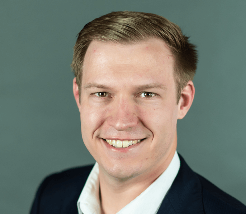A well-dressed man in a suit and tie smiles warmly for a photograph, showcasing a friendly and professional demeanor.